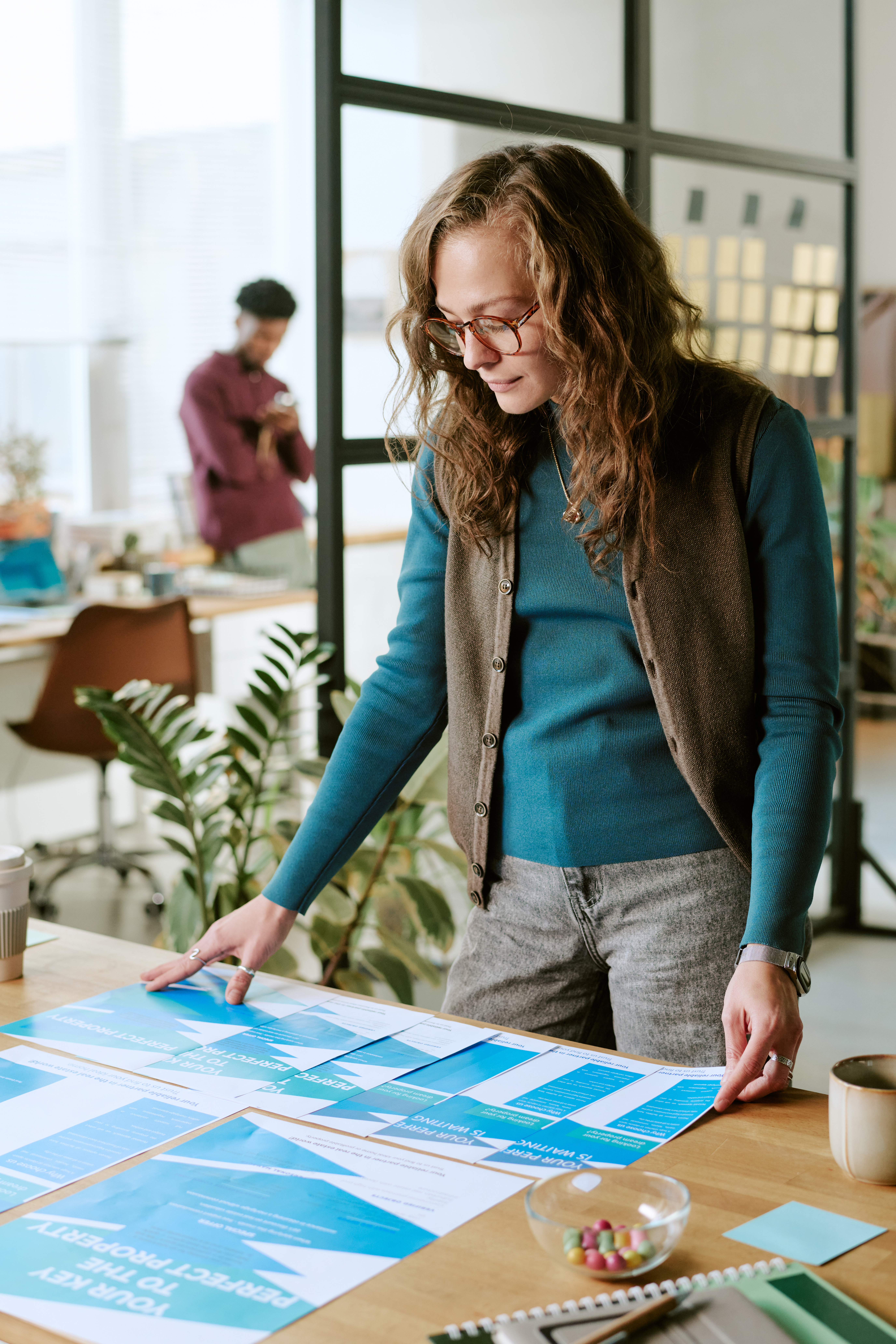 Caucasian young adult woman standing at desk reviewing printed charts and documents, wearing eyeglasses, working in modern office with blurred male colleague in background