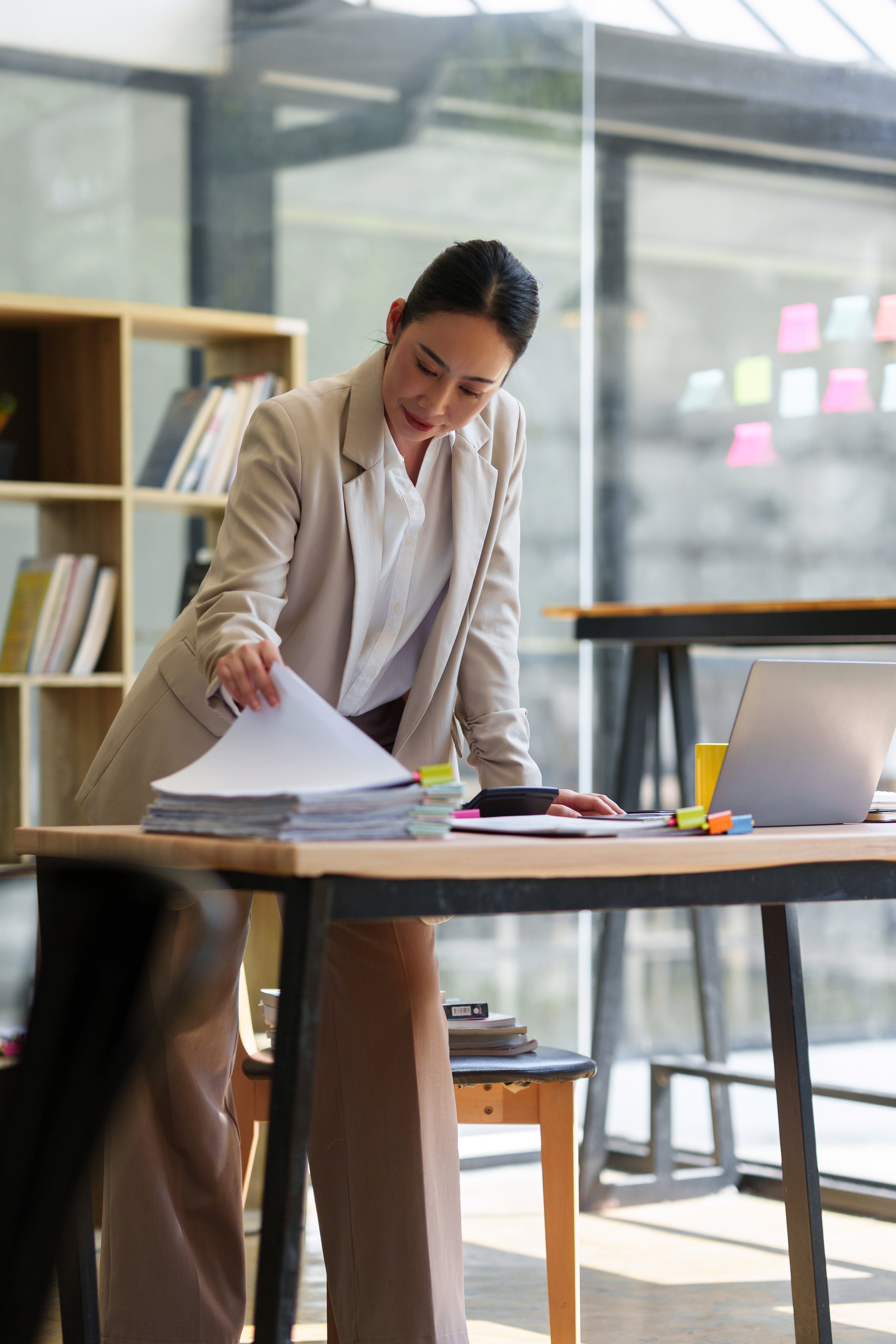 Professional businesswoman standing at an office desk, organizing a large stack of documents and paperwork with a laptop nearby